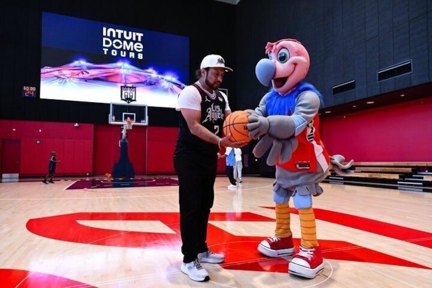 Ein Maskottchen hält einen Basketball auf einem Spielfeld, im Hintergrund eine große Leinwand mit der Aufschrift 'INTUIT DOME TOURS'. 5