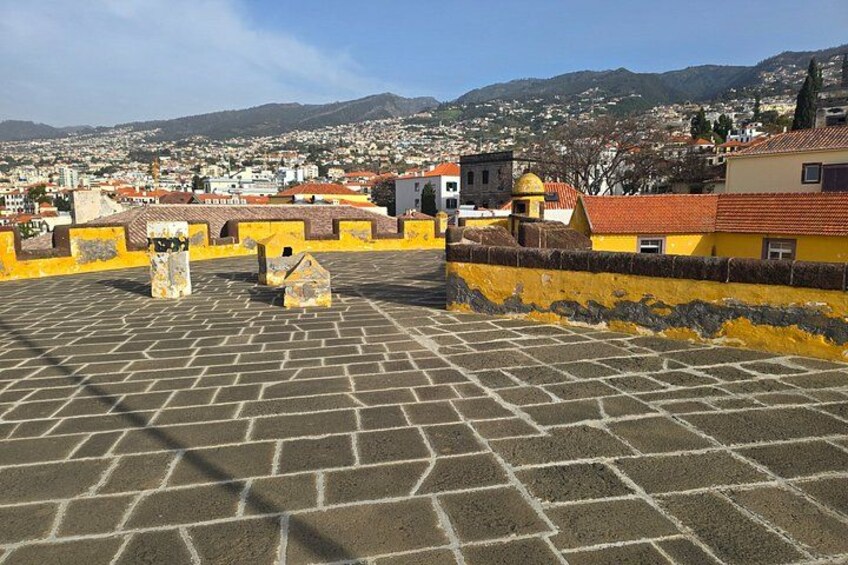 A historic fortification with yellow walls and a paved courtyard, overlooking a cityscape with buildings and hills in the background. 4