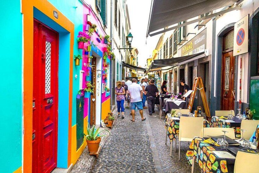 A narrow street with colorful buildings, outdoor seating, and people walking. 1