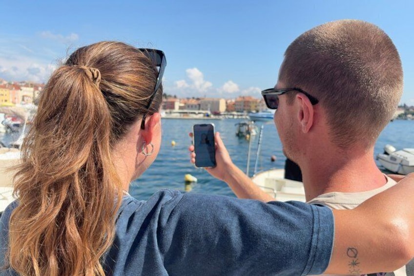 A person taking a photo with a smartphone, another person with a tattoo on their arm, and a body of water with boats in the background. 1