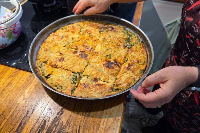 Iemand houdt een ronde, gebakken schotel met een goudbruine korst vast, mogelijk een quiche of frittata, op een houten tafel. 3