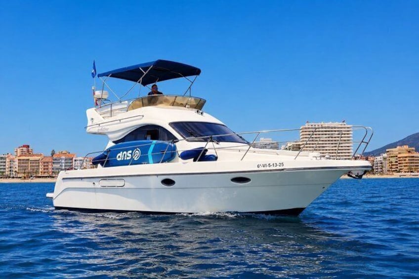 Uno yacht bianco con marchio 'dns' sul fianco, che naviga in acque calme sotto un cielo sereno con una città costiera sullo sfondo. 2