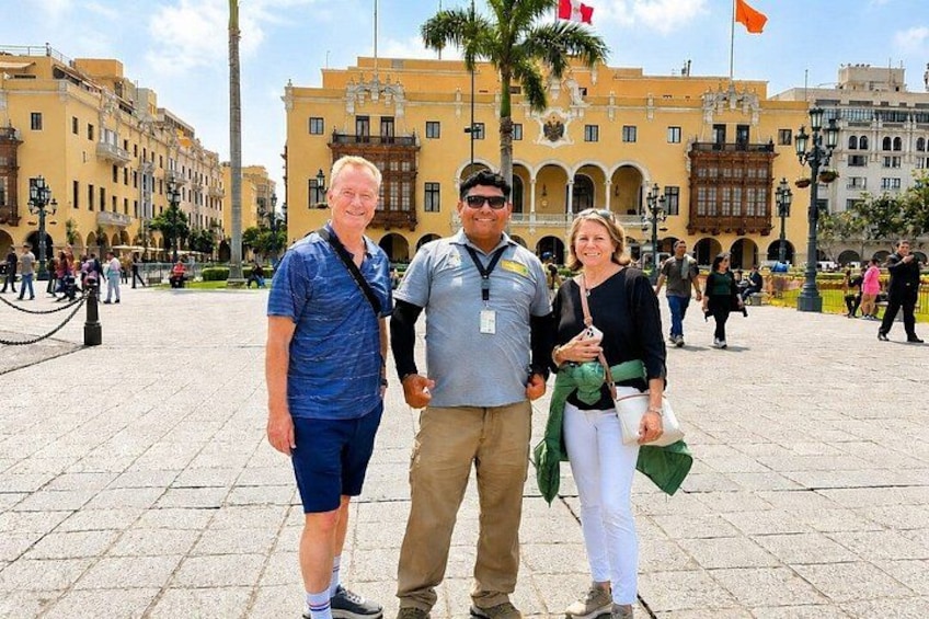 Tres personas de pie en una plaza pública con edificios históricos al fondo. 1