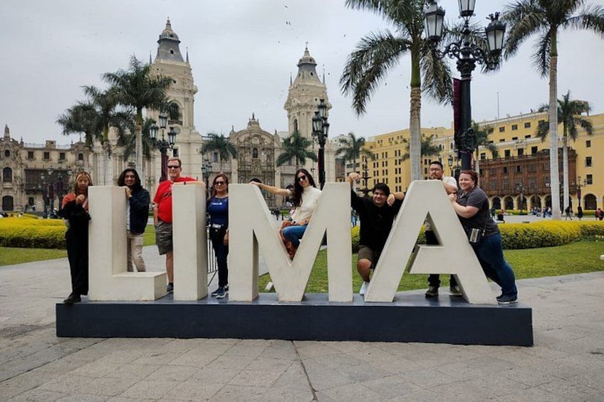 Un grupo de personas frente a un letrero grande con la palabra 'LIMA', con un edificio histórico y palmeras de fondo. 3