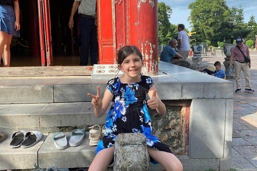 Un enfant assis sur un banc de pierre, levant le pouce en signe d’approbation, entouré d’une colonne rouge, d’un mur de pierre et d’un sol carrelé. 2