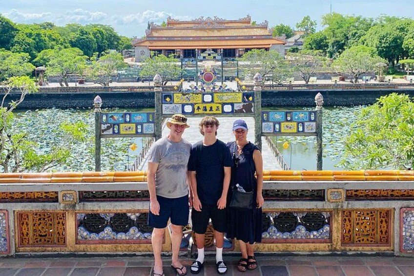 A group of people standing on a bridge with a decorative railing, a body of water, and a traditional building in the background. 2