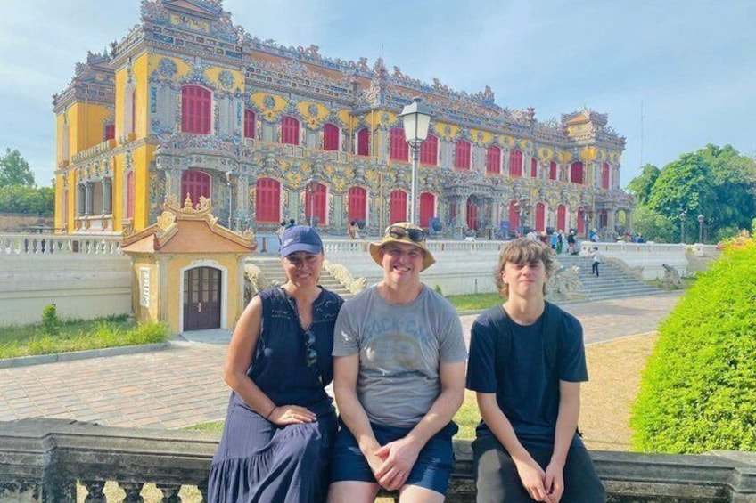 Three people sitting on a bench in front of a large, ornate building with red windows and a yellow facade. 4
