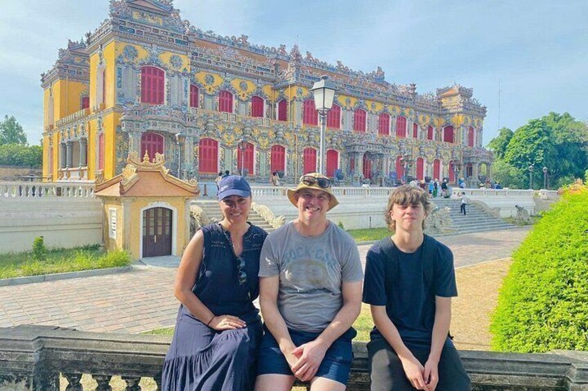 Un groupe de personnes est assis sur un banc devant un grand bâtiment orné, dont les couleurs dominantes sont le rouge et le jaune. 3