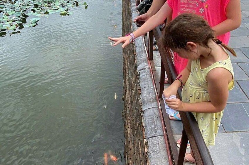 Ein junges Mädchen in einem gelben Kleid steht an einem Geländer und berührt das Wasser. Im Hintergrund ein Teich mit Seerosenblättern. 1