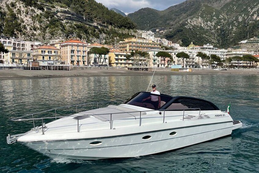 Un bateau à moteur blanc, doté d’une cabine, navigue sur une eau calme, avec une ville côtière et des montagnes en arrière-plan. 4