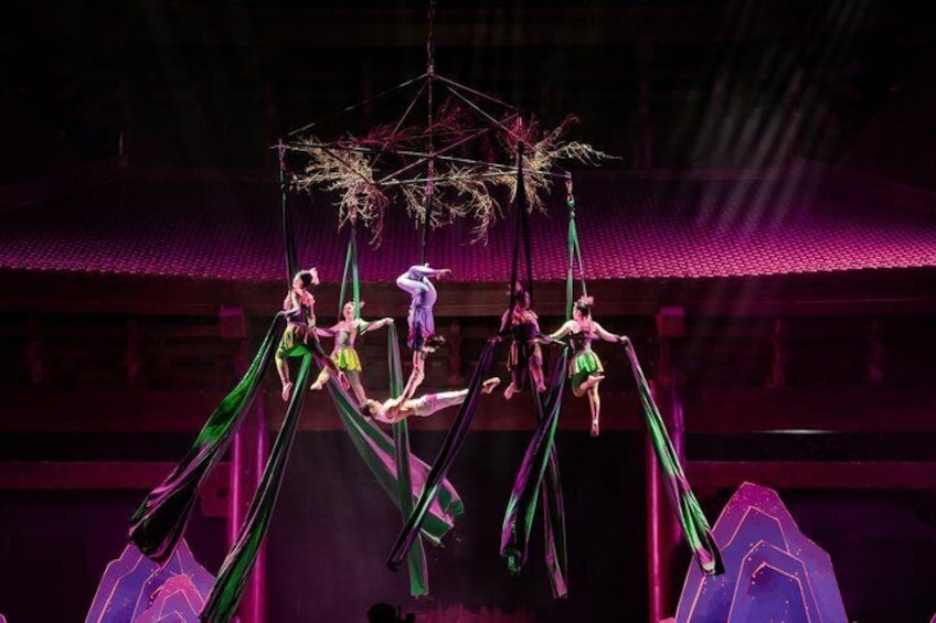 Aerial performers in green costumes performing on a suspended structure with a traditional building in the background. 1