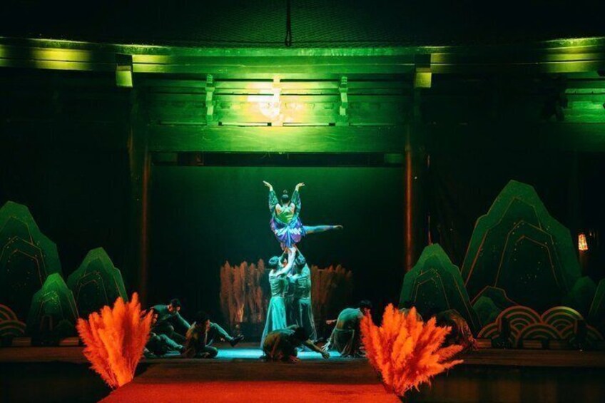 A stage with a performer in traditional attire, a backdrop with a mountain design, and red foliage on the floor. 3