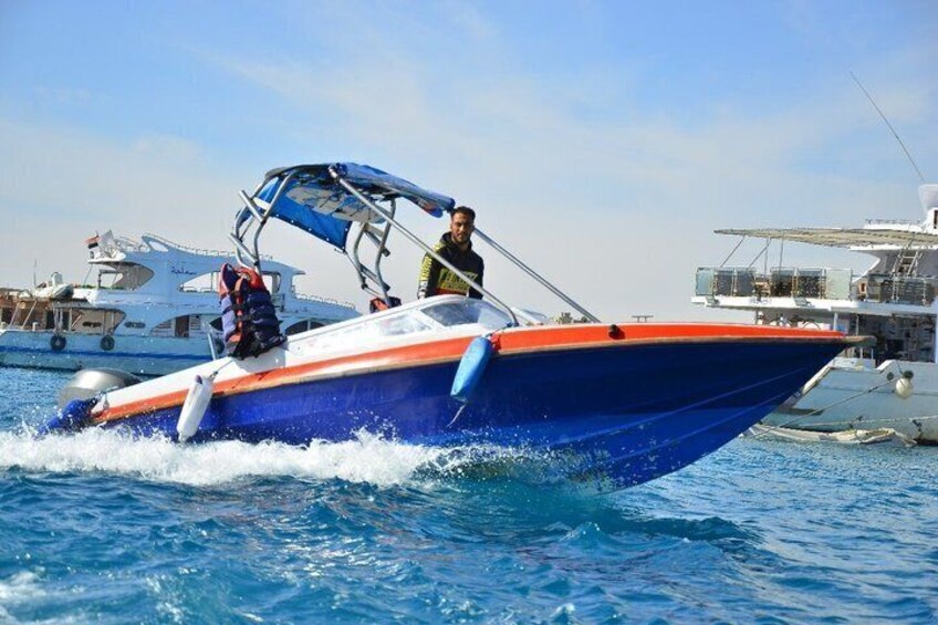 Een blauw en oranje speedboot met een kap, vaart door rustig water, met andere boten op de achtergrond. 1