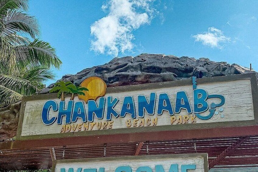 Señal para el Parque de Aventuras Playa Chankanaab, con palmeras y montañas al fondo. 1
