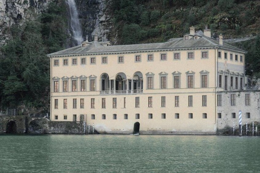 Un grande edificio a più piani con finestre ad arco e un balcone, affacciato su uno specchio d'acqua con una cascata sullo sfondo. 3