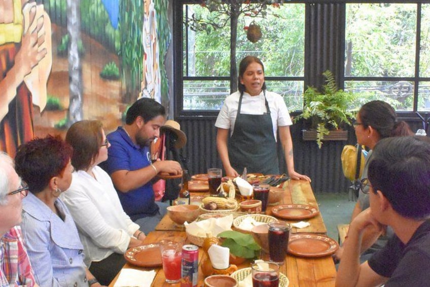 Un grupo de personas sentadas alrededor de una mesa de madera con varios platos y bebidas, una persona parada al frente sirviendo comida, y un mural en la pared de fondo. 1