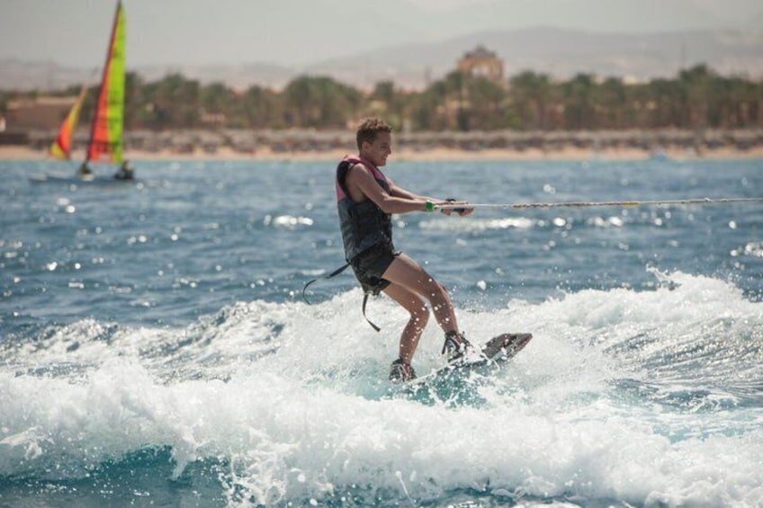 Een persoon wakeboard achter een wateroppervlak met golven, een zeilboot op de achtergrond, en een kustlandschap met gebouwen. 5