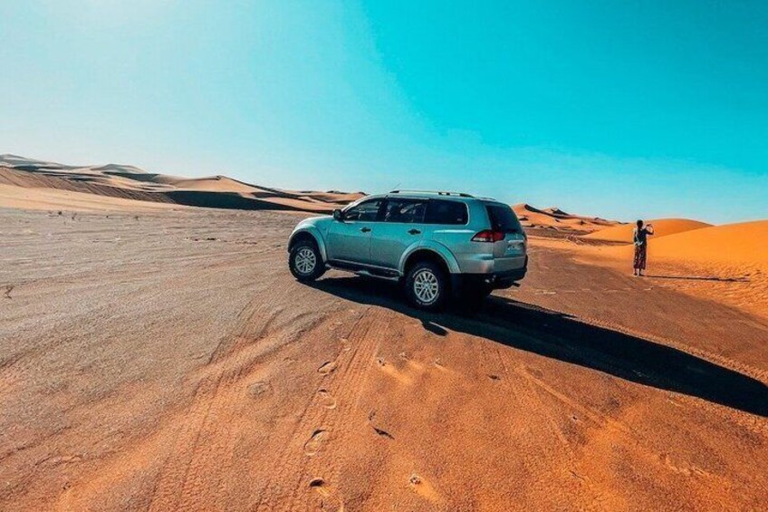 A light blue SUV parked on a desert road with sand dunes in the background. 1