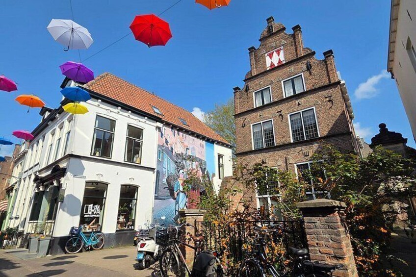 A street with colorful umbrellas, a white building with a mural, and a brick building with a red and white sign. 1