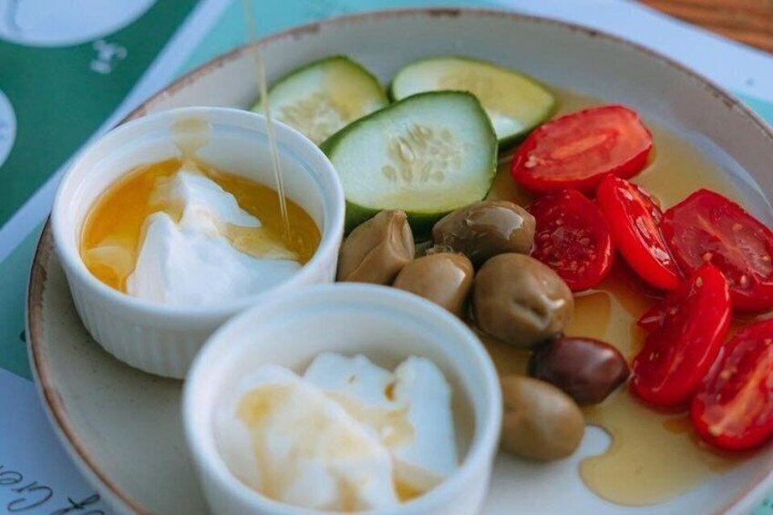 A plate with sliced cucumbers, cherry tomatoes, olives, and two bowls of a creamy white substance, possibly yogurt or sour cream. 2