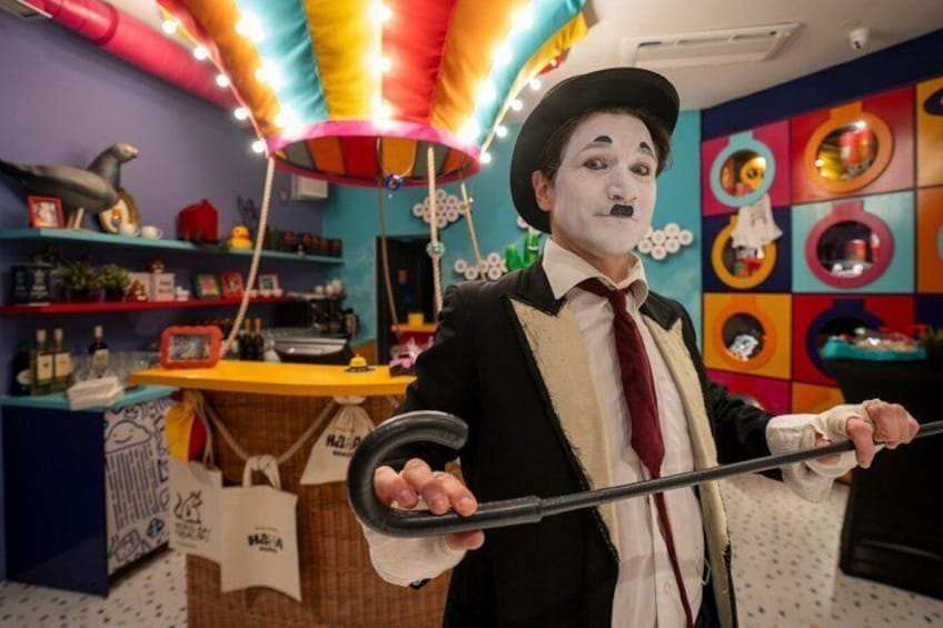 Une personne vêtue d’un costume et d’un chapeau noirs, tenant une canne, se trouve dans une pièce colorée, dotée d’étagères et d’un comptoir. 5