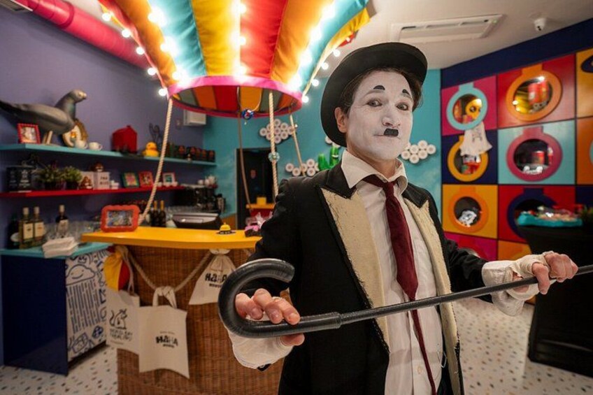 Une personne vêtue d’un costume et d’un chapeau noirs, tenant une canne, se tient dans une pièce colorée, où l’on aperçoit des étagères et un carrousel. 5