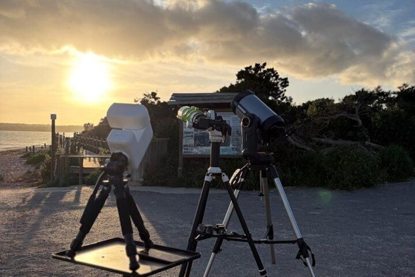 Un télescope monté sur un trépied, une personne debout sur une plateforme et un coucher de soleil sur une plage. 1