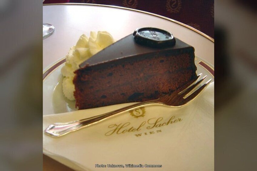 A slice of chocolate cake on a plate with whipped cream, a fork, and a napkin with 'Hotel Sacher' written on it. 3