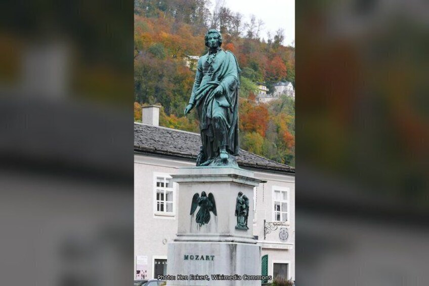 Une statue représentant un homme tenant un enfant, placée sur un piédestal, avec en arrière-plan des arbres aux couleurs automnales et un bâtiment. 3
