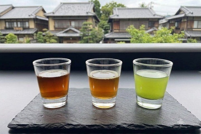 Tres vasos de líquido sobre un posavasos de pizarra, con edificios tradicionales japoneses al fondo. 4