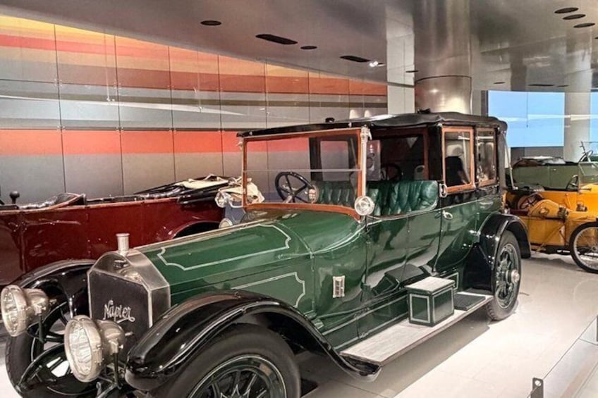 En klassisk grön bil med svart tak och träinredning, utställd i en modern byggnad med reflekterande väggar. 5