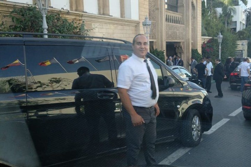 A man standing next to a black vehicle with flags on its side, a building with columns, and people in the background. 4