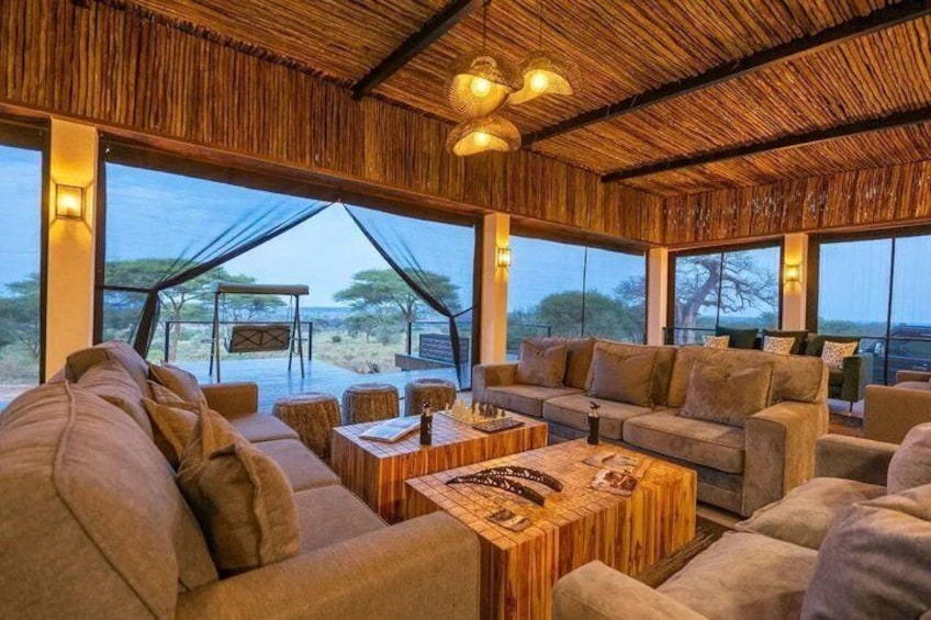 An indoor lounge with sofas, wooden coffee tables, and large windows overlooking a savannah landscape. 3