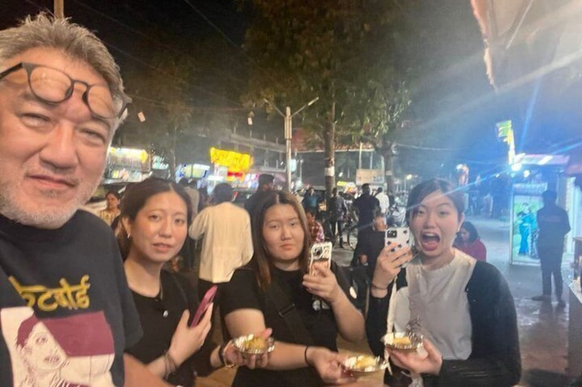 A group of people holding food items, standing on a street with shops and trees in the background. 2