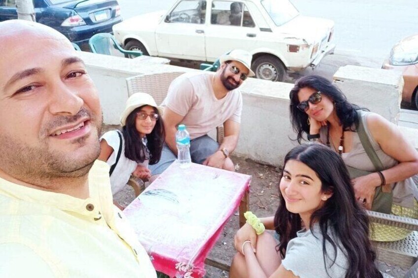 A group of people sitting at an outdoor table with a pink tablecloth, parked cars in the background, and a concrete barrier. 2