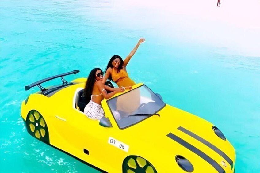 Dos personas en un coche descapotable amarillo sobre una masa de agua turquesa. 1