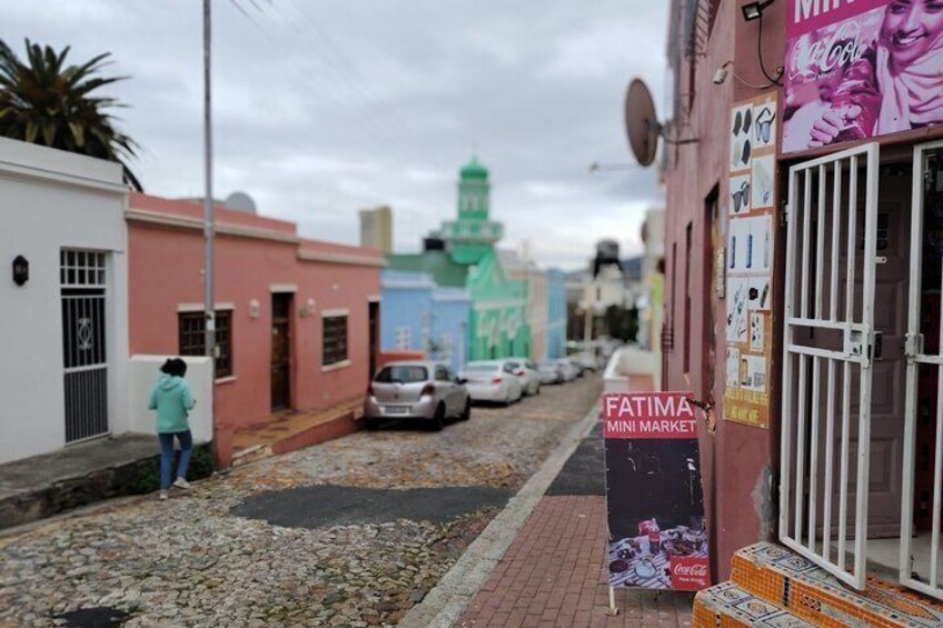 Una strada con edifici colorati, un'auto parcheggiata e una persona appoggiata a un muro. 3