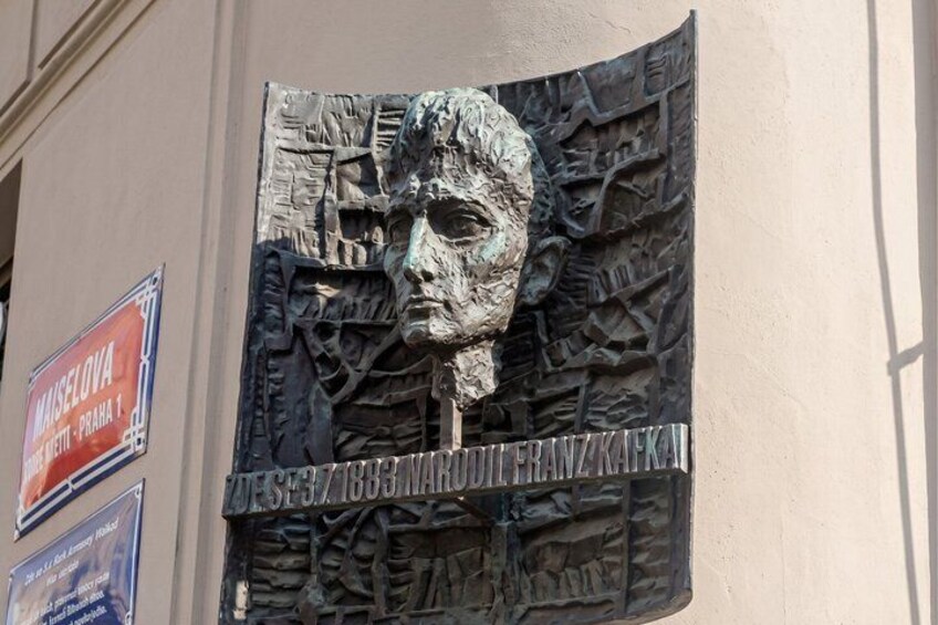 A sculpture of a man's face with a bust of a person below it, and a sign with the text 'MAZELOVA' and 'DESEK. 3. 1. 1835. NARODNIKAFKA.' 3
