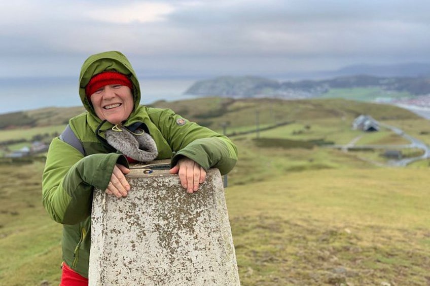 A person leaning on a weathered stone marker, wearing a green jacket and red cap, with a hilly landscape and a distant road in the background. 2