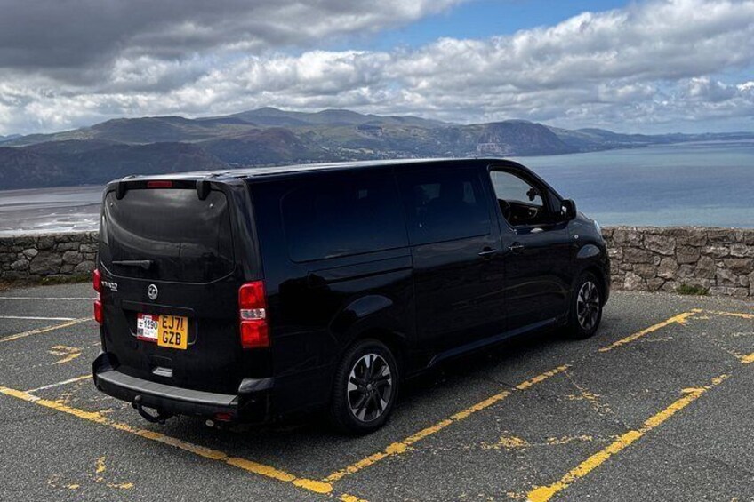 A black van parked in a parking lot with a scenic view of mountains and a body of water in the background. 4