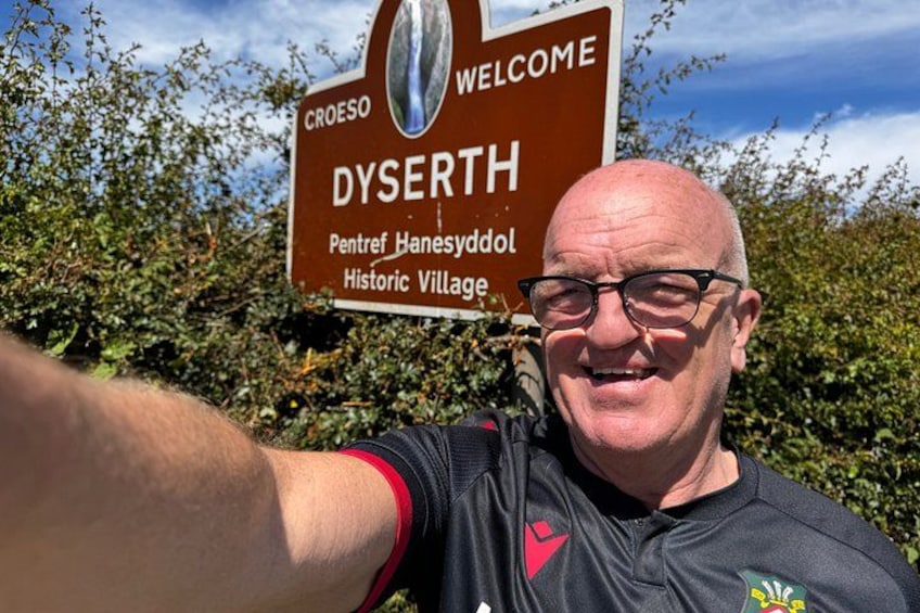 A man taking a selfie with a sign reading 'Welcome to Dysherth, Pentre Hanesyddol, Historic Village.' 4