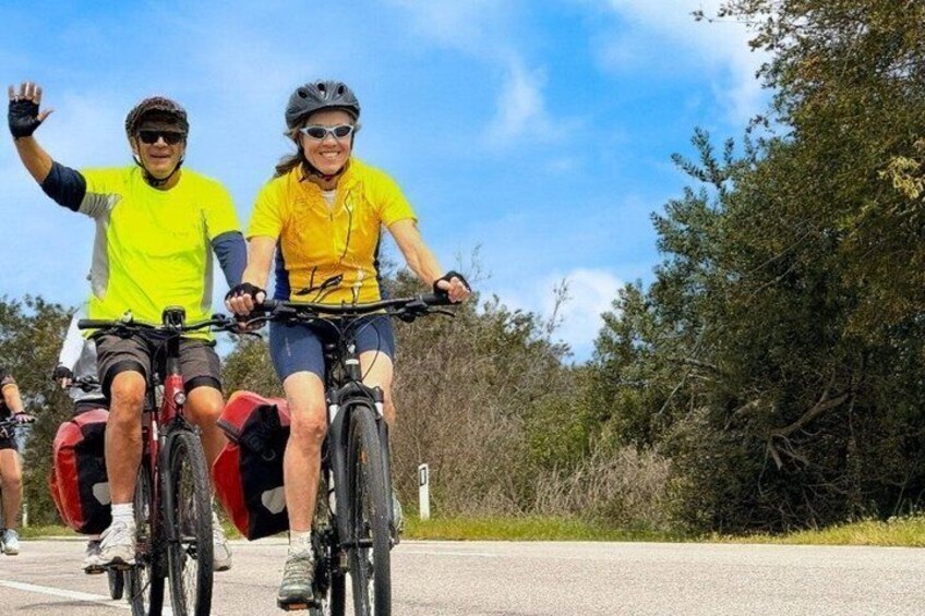 Two people riding bicycles on a paved path with trees and a clear sky in the background. 2