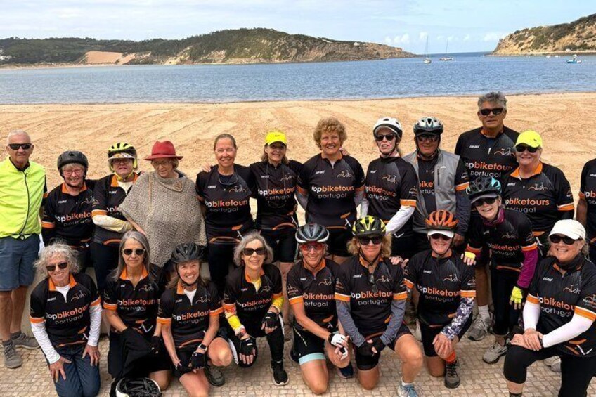 A group of people in cycling attire posing on a beach with a body of water and hills in the background. 1