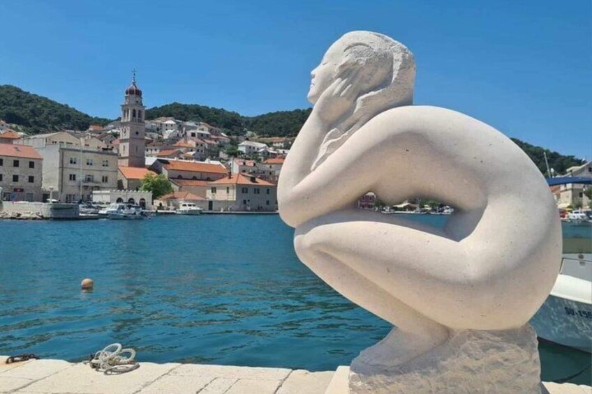 Una escultura de una figura pensativa junto al agua, con un pueblo y una iglesia al fondo. 3