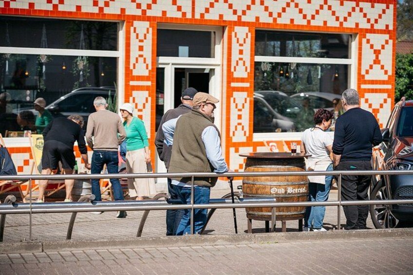 A group of people gathered outside a building with a distinctive orange and white pattern, a wooden barrel labeled 'Old Beer,' and a bicycle. 2