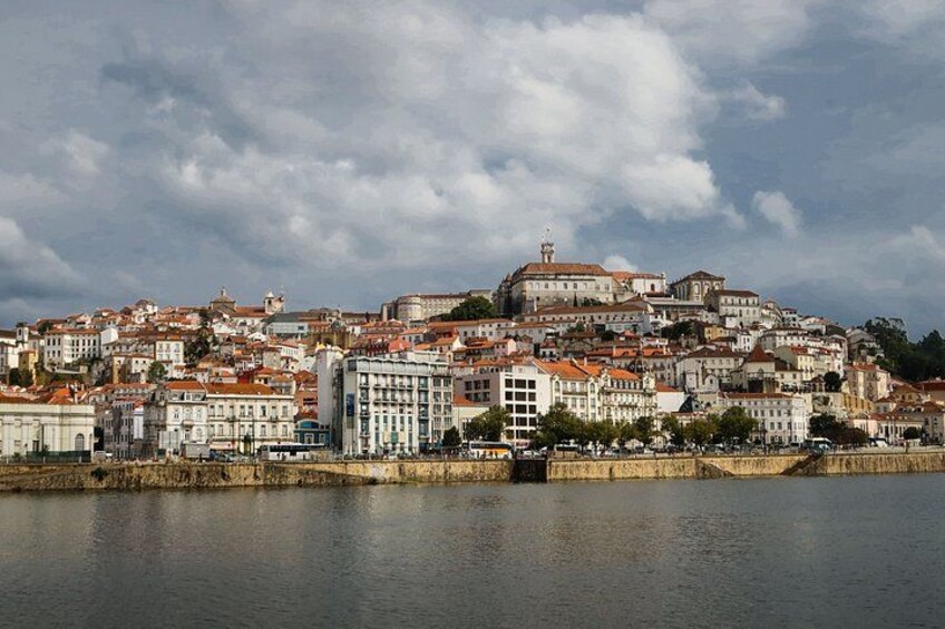 A coastal cityscape with historic buildings, a prominent hilltop structure, and a calm body of water. 1