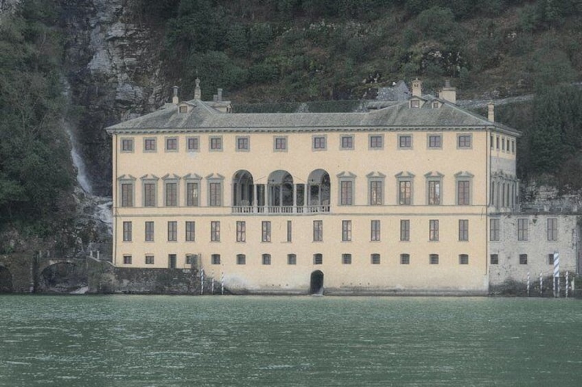A large, two-story building with a symmetrical facade, arched windows, and a central entrance, situated by a body of water with a rocky cliff in the background. 3