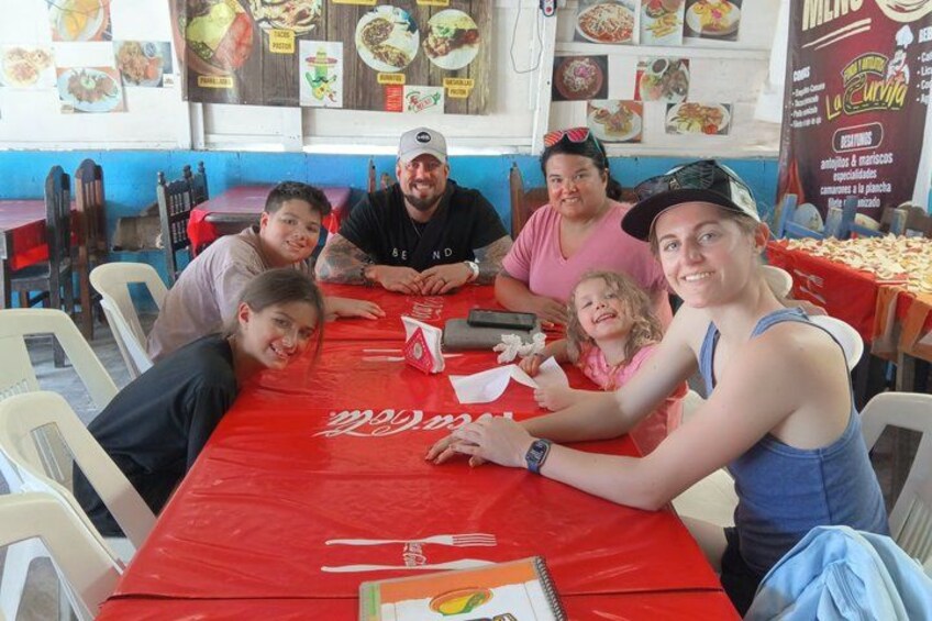 Un groupe de personnes est assis autour d’une table rouge ornée du logo Coca-Cola, dans un restaurant où l’on aperçoit, au fond, des panneaux affichant le menu. 5