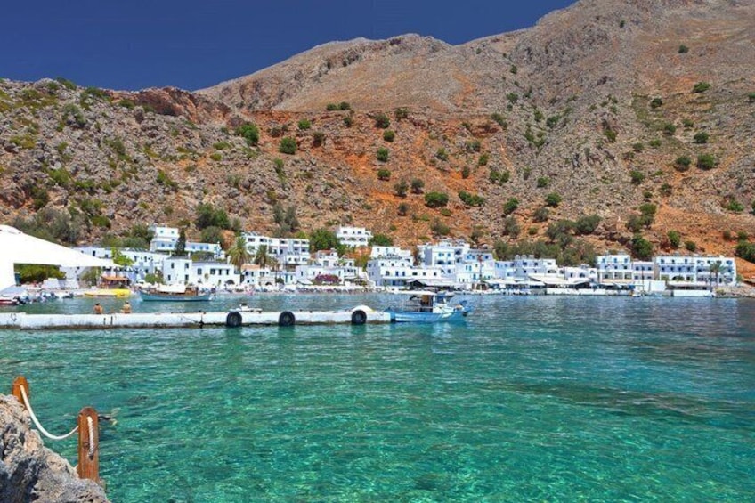 A coastal village with white buildings, clear turquoise waters, and a mountainous backdrop. 1
