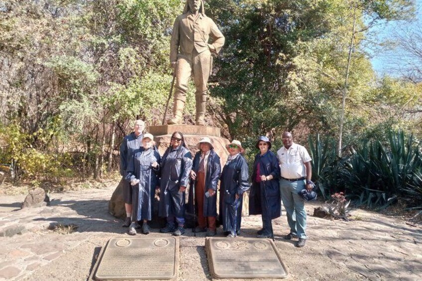 Eine Gruppe von Menschen steht vor einer Statue in einem Park mit Bäumen und einem Pflasterweg. 2